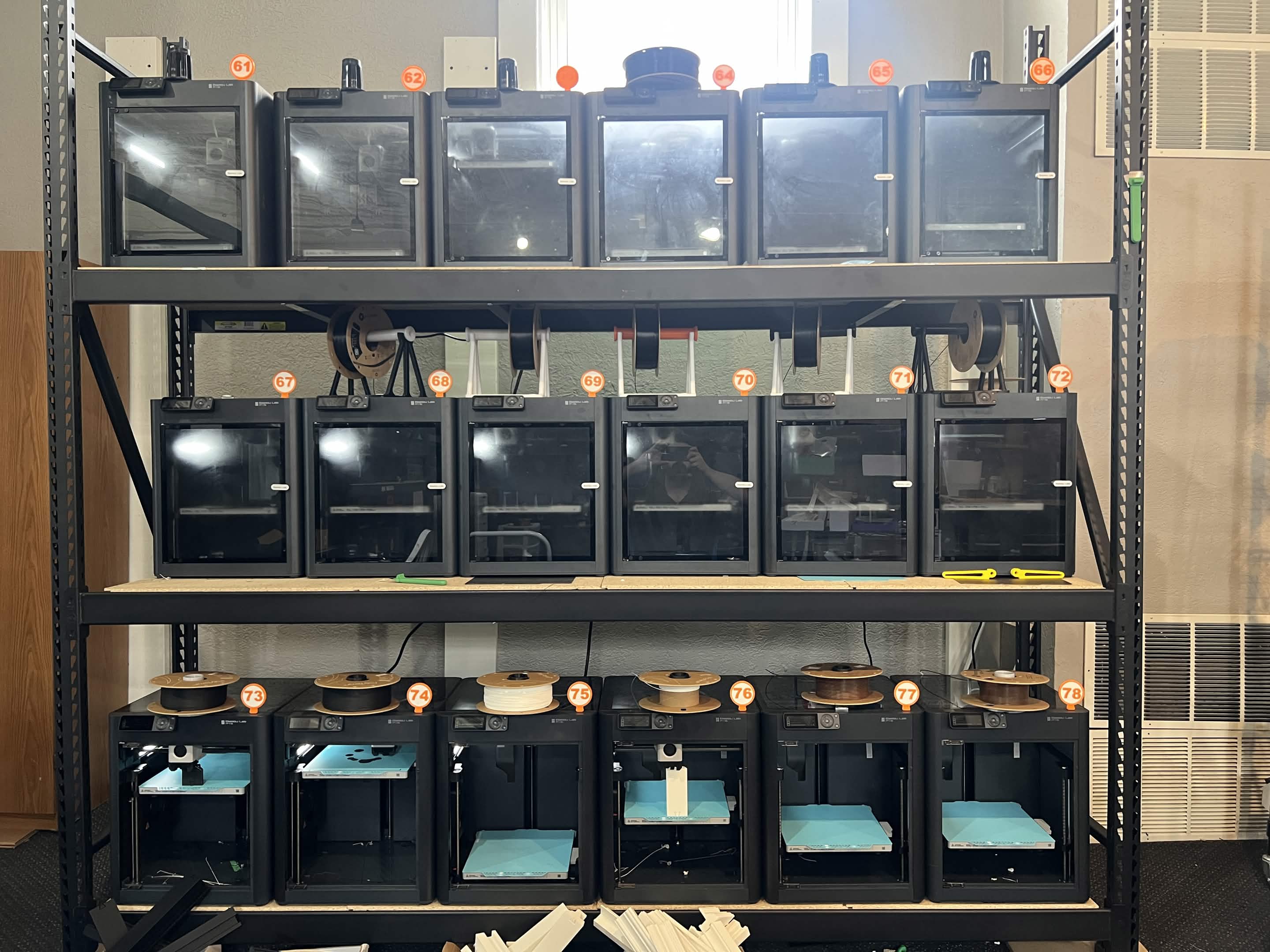 Front view of multiple enclosed 3D printers arranged across several shelves in the GoodPrints3D farm, reinforcing real small-batch and repeat-order capacity.