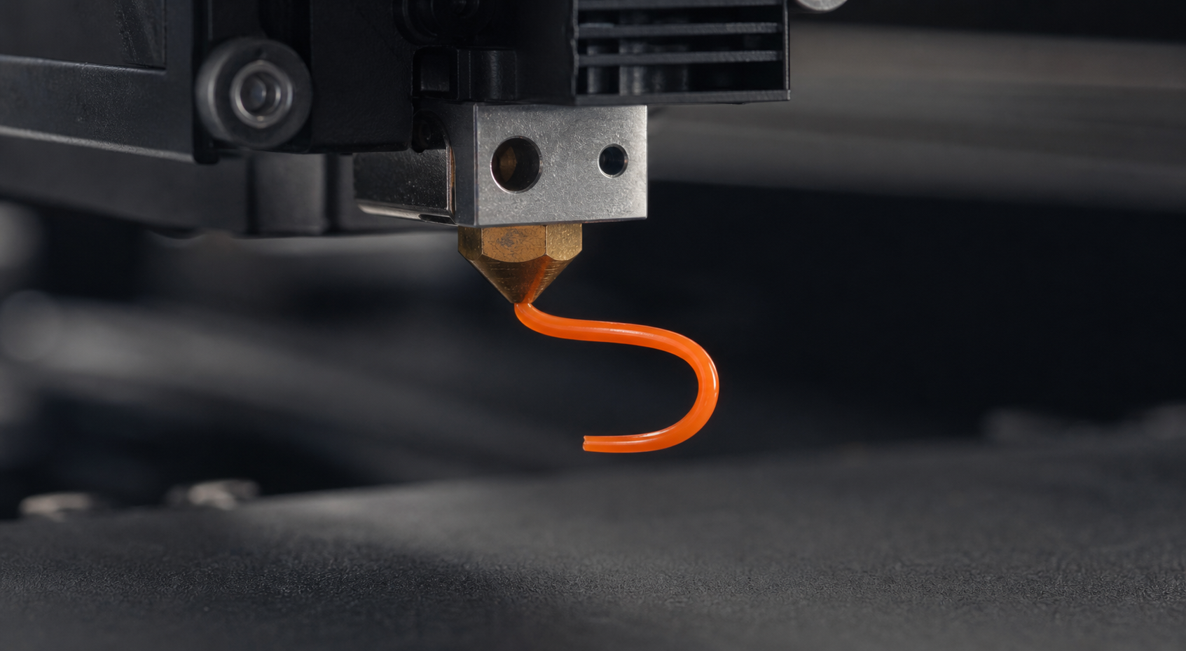 Macro view of orange filament curling sharply sideways as it is extruded from a brass 3D printer nozzle.