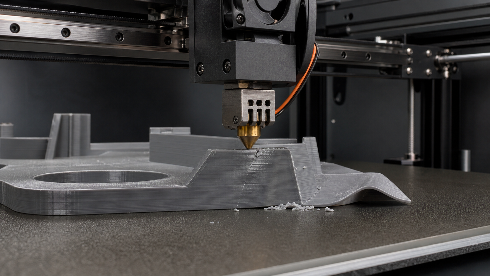 Close-up of a brass 3D printer nozzle scraping a raised gray print surface, with a curled edge and small plastic crumbs visible on the build plate.